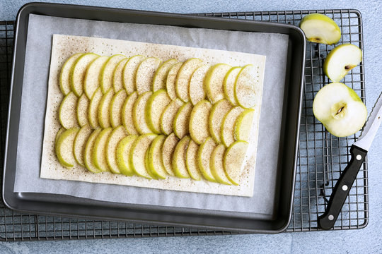 An uncooked rectangular tart with rows of apple on the pastry, a knife and cut pieces of cooking apple.