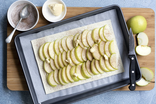 An uncooked apple tart with rows of sliced green apples on pastry, a sprinkling of cinnamon sugar and dots of butter.