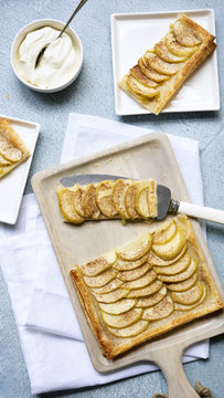 A rectangular apple tart on a wooden serving board cut in pieces being served onto dessert plates.