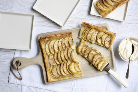 An apple tart cut into pieces on a wooden serving board, with plates and a bowl of cream.