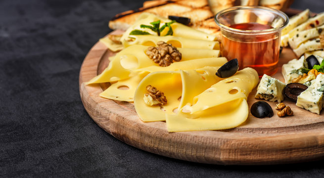 Cheese plate: Parmesan, cheddar, gouda, mozzarella and other with basil on wooden board on dark background with place for text.Honey and Crackers