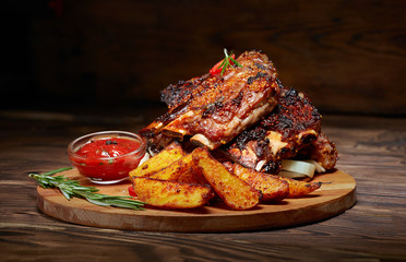 Fried ribs with rosemary, potatoes rustic, onion, sauce on wooden round Board. Dark background....