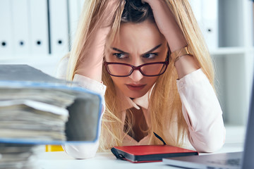 Tired and exhausted woman in spectacles looks at the mountain of documents propping up her head...