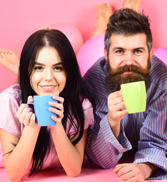 Couple relax in morning with coffee. Family tradition concept. Man and woman on smiling faces lay, pink background. Couple in love drink coffee in bed. Man and woman in domestic clothes, pajamas.