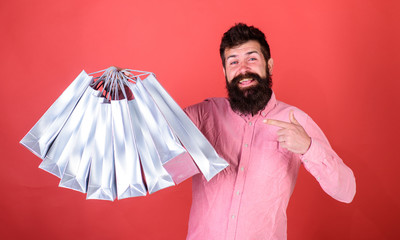Man with beard and mustache pointing at shopping bags, red background. Hipster on happy face is...