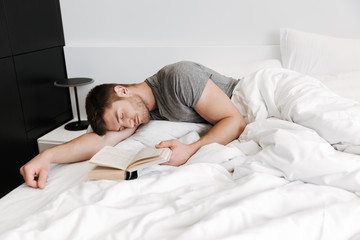 Attractive young man sleeping in bed while holding open book