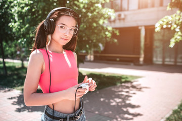 Attractive and amazing girl is looking at camera and posing. She is listening to music in headphones. Also girl is holding white player in her hands. She is standing on the street.