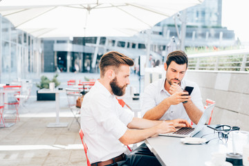 Obraz premium two bearded young business young men outdoor using laptop and smart phone remote working - finance, trading online, wifi technology concept