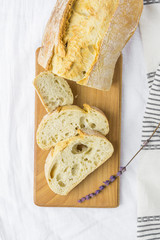 Top View of Sliced Artisan Rustic Bread on Wooden Cutting Board White Line Table Cloth Lavender Twig. Golden Crust Appetizing Spongy Holey Texture. Mediterranean Cuisine Specialty. Provence Style