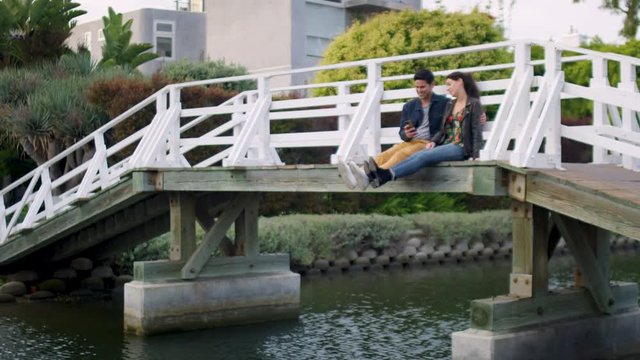 Cute Couple Sit On Bridge In Venice Beach Canals And Take Fun Photos Of Their Feet Above Water (Slow Motion)