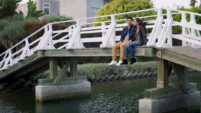 Couple Sit On Edge Of Bridge And Enjoy View Of Venice Beach Canals Together, Man Puts His Arm Around Girlfriend (Slow Motion)
