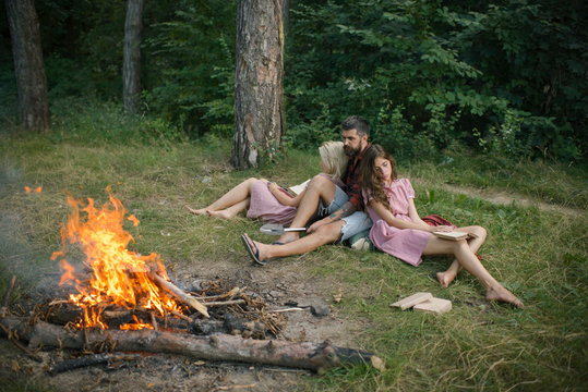 Friends relax at campfire. People at bonfire flame in green forest. Women and man at fire in camp. Camping, hiking and travel. Summer vacation concept