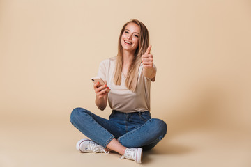 Obraz na płótnie Canvas Image of happy young woman wearing casual clothing showing thumb up and holding mobile phone while sitting on floor with legs crossed, isolated over beige background