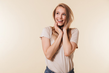 Portrait of gorgeous woman 20s with european appearance in basic t-shirt smiling and looking upward touching her hair, isolated over beige background in studio