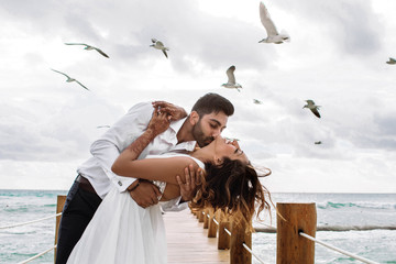 Seagulls fly over gorgeous wedding couple kissing on the wooden quay over the sea