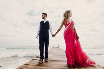 Wind blows bride's pink sari while she stands with Hindu groom on a wooden quay among foaming waves