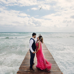 Wind blows bride's pink sari while she stands with Hindu groom on a wooden quay among foaming waves
