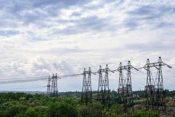 High voltage lines and power pylons in a green forest, sunny day with clouds in the sky. in the background building, city, village, power supply, electrification, electricity transmission problems