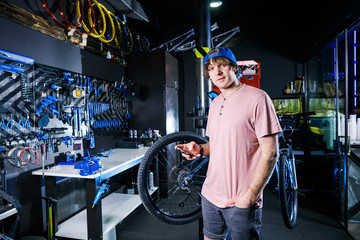 A young, stylish redheaded male small business owner selling and repairing a bike is wearing a blue cap and a pink jersey is using a mobile phone while standing in a bike shop