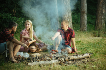 Group of friends camping in forest. People having fun talking around campfire. Cooking sausages...