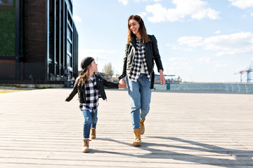 Full-lenght outside portrait of young pretty woman with little adorable girl walking in sunlight near the ocean dressed in similar outfits with happy smiles