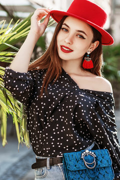 Outdoor portrait of young beautiful fashionable happy smiling girl wearing stylish red hat, earrings, polka dot blouse, blue waist bag posing in street. Summer fashion concept