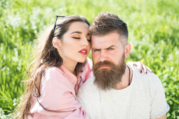 Couple in love on green grassy meadow. Sensual brunette in trendy eyewear embracing brutal bearded man. Sexy girl with full red lips hugging frowning macho on flowery field, spring time romance