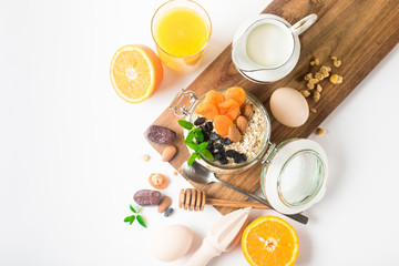 Healthy breakfast in glass mason jar with oat flakes, granola, milk, dry fruits and nuts, dates, honey, orange juice, eggs for morning fitness on wooden board platter on white background isolated