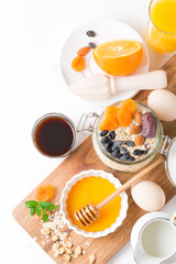 Healthy breakfast in glass mason jar with oat flakes, granola, milk, dry fruits and nuts, dates, honey, orange juice, eggs for morning fitness on wooden board platter on white background isolated