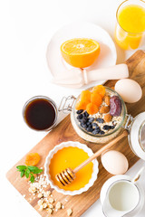 Healthy breakfast in glass mason jar with oat flakes, granola, milk, dry fruits and nuts, dates, honey, orange juice, eggs for morning fitness on wooden board platter on white background isolated