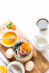 Healthy breakfast in glass mason jar with oat flakes, granola, milk, dry fruits and nuts, dates, honey, orange juice, eggs for morning fitness on wooden board platter on white background isolated