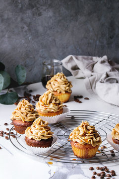 Fresh baked homemade cupcakes with coffee buttercream and caramel standing on cooling rack with eucalyptus branch and coffee beans above over white marble kitchen table.