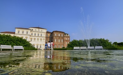 Fototapeta na wymiar Venaria reale, Piedmont region, Italy. June 2017. The facade of the palace, on the side of the gardens, is reflected on the fountain's water mirror. Tourists explore the magnificent park.
