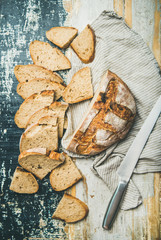 Flat-lay of freshly-baked sourdough wheat bread loaf halved and cut in slices over linen napkin and rustic table background, top view, vertical