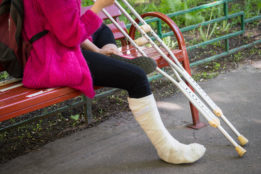 unrecognizable girl sits on the street on a bench with a broken leg and crutches