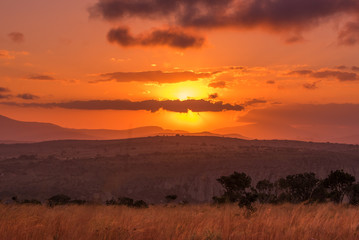 The sun glows behind clouds in an intense sunset above an African landscape of hills, cliffside and...