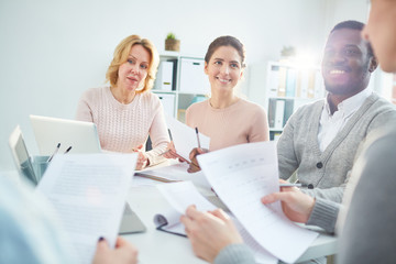 Cheerful multi-ethnic team of managers analyzing results of accomplished work with help of statistic data while gathered together at boardroom illuminated with sunbeams