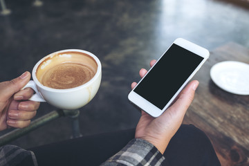 Mockup image of a woman's hand holding white mobile phone with blank black desktop screen and coffee cup in cafe