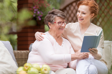 Cheerful senior woman with her tender caretaker reading a book together while relaxing outside....