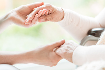 Close-up of a young woman and a senior lady holding hands with tenderness and care. Showing support and love. Blurred background.