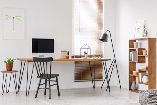 Black chair at wooden desk with computer monitor in bright home office interior with lamp. Real photo