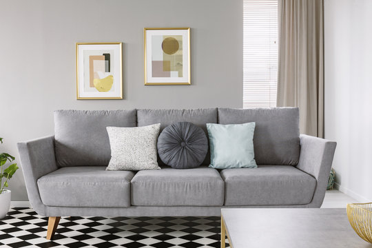 Real photo of a grey sofa with black and white pillows in a living room interior with checkered floor and posters in gold frames on a wall