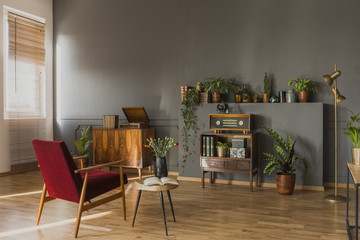 Dark red armchair next to table with flowers in grey vintage flat interior with radio on cabinet. Real photo