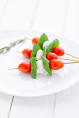 cherry tomatoes with basil and mozzarella cheese on .wooden skewers placed on a white plate and white wooden table