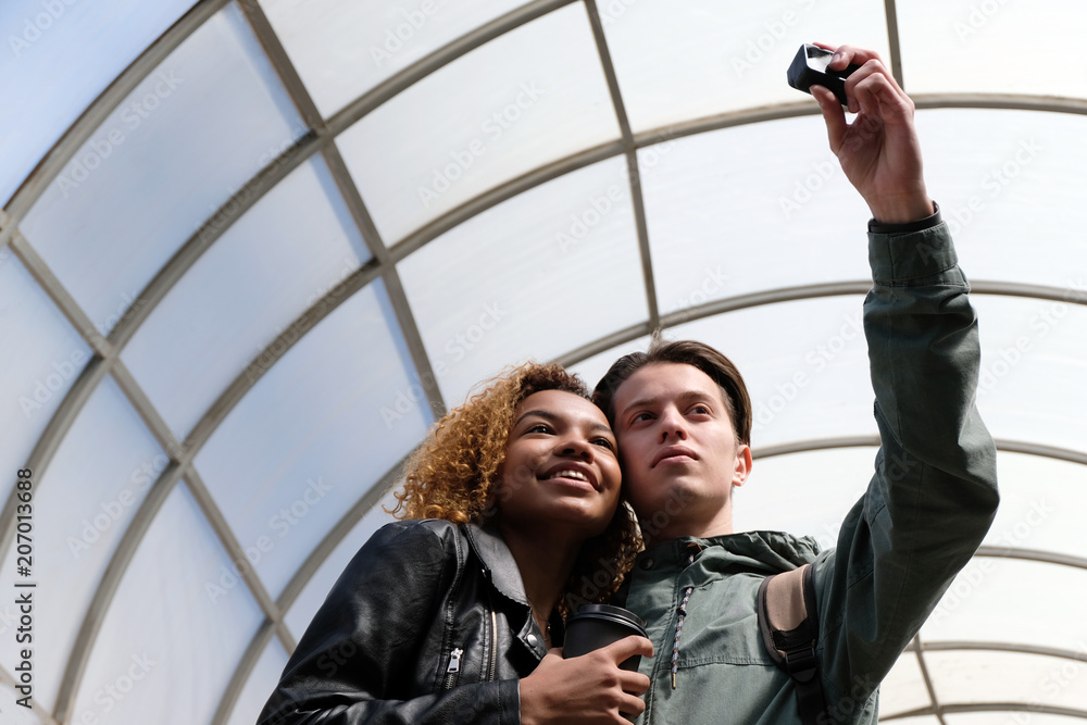 Wall mural A modern beautiful African American girl smiles when together with a white guy they make selfi on an action camera. Cute international friends-students are photographed together inside a glass dome.