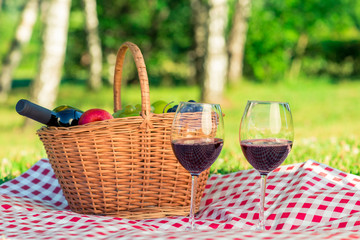 basket with red wine and fruits and two glasses of wine on a tablecloth on a lawn in a park, close-up