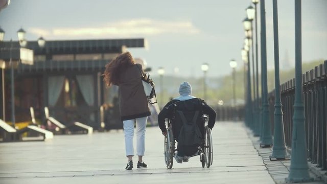 Young happy woman with disabled man in a wheelchair walking together on the quay