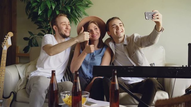 Playful young people from musical band are taking selfie with smartphone, posing and making funny faces, laughing and hugging. Musical instruments are visible.