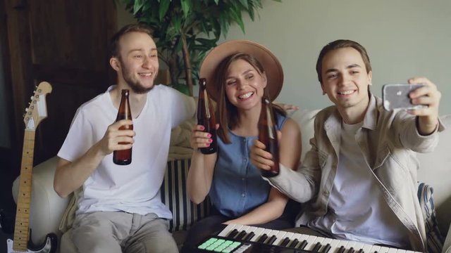 Happy musicians are taking selfie with beer bottles sitting on sofa in home studio and holding smartphone. Young people are posing, clanging bottles and having fun.