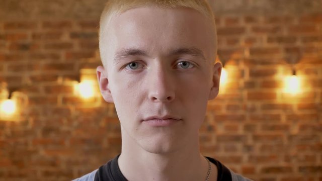 Young serious man hipster watching at camera, brick background, close up